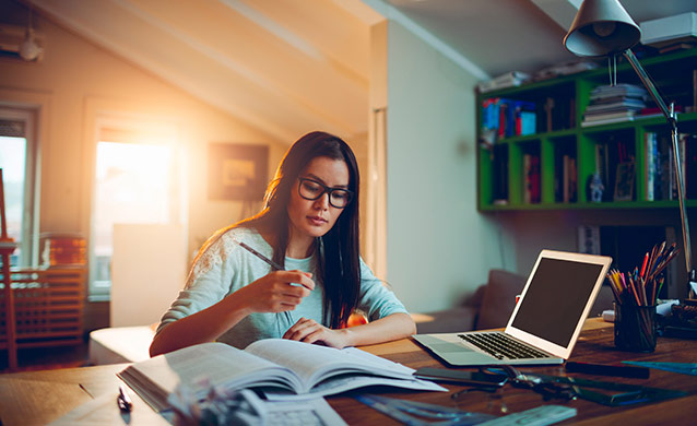 student studying at desk in her home with a book and a laptop